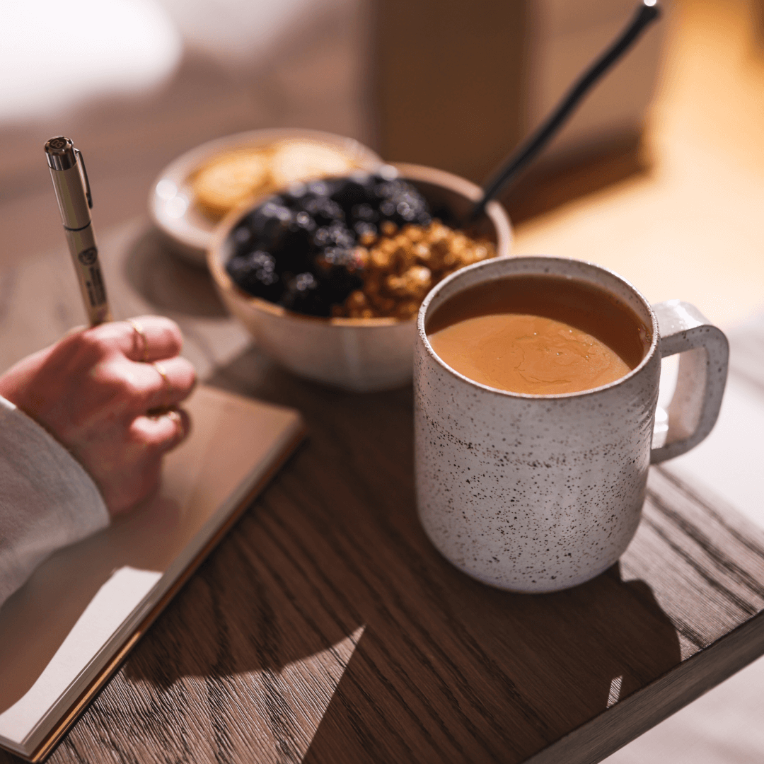 Person writing in a notebook with coffee and a bowl of berries and granola nearby.